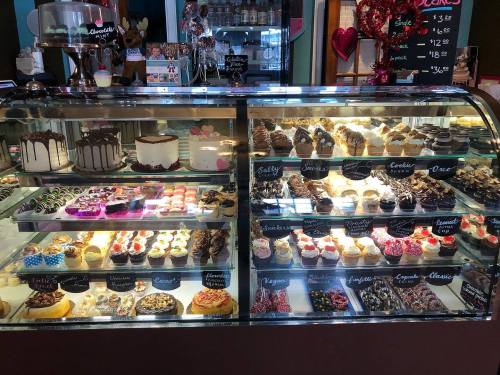A display case filled with various cakes, cupcakes, and desserts, showcasing colorful treats and decorative labels.