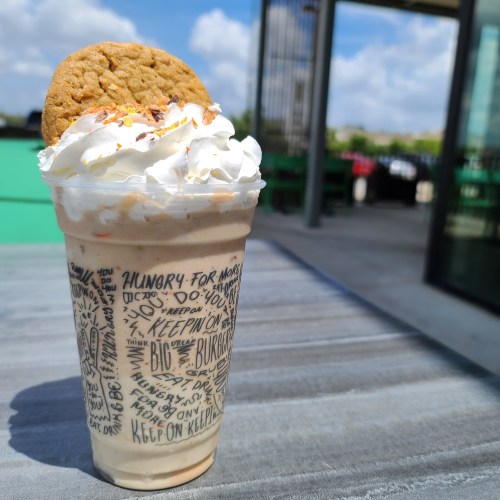 A creamy milkshake topped with whipped cream and a cookie, served in a decorated cup on a sunny outdoor table.