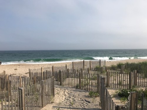 A serene beach scene with gentle waves, sand dunes, and wooden fencing under a cloudy sky.