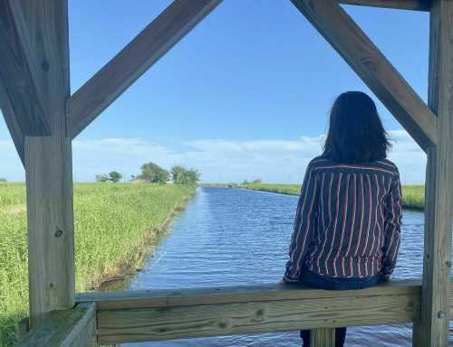 A person sits on a wooden structure, gazing at a calm waterway surrounded by green grass and blue sky.