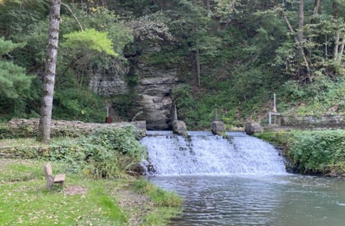 A serene scene of a small waterfall cascading over rocks, surrounded by lush greenery and a bench nearby.