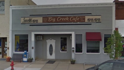 Exterior of Big Creek Cafe, a gray building with a sign, white door, and red awning, located in a small town.