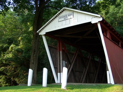 A historic covered bridge with a white roof and red walls, surrounded by lush green trees.