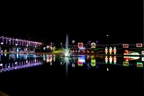 Colorful holiday lights reflect on a calm water surface at night, featuring festive decorations and a fountain.