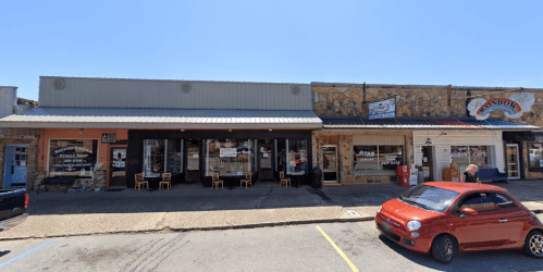 A row of small shops with outdoor seating, featuring a red car parked in front on a sunny day.