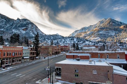 Snow-covered mountains loom over a quiet town street lined with historic buildings under a dramatic sky.
