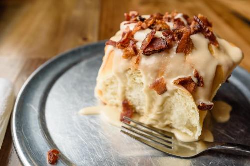 A slice of sweet roll topped with creamy frosting and crispy bacon bits on a metal plate.
