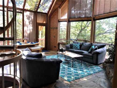 Cozy living room with large windows, spiral staircase, and comfortable seating, surrounded by nature.
