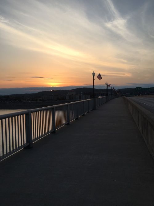 A peaceful walkway along a river at sunset, with flags and soft clouds in the sky.