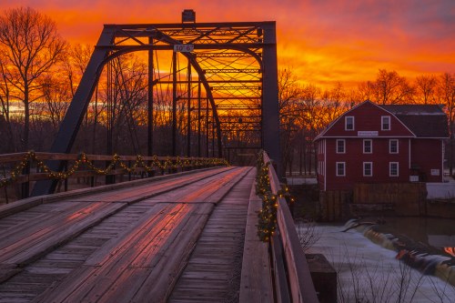 A historic bridge at sunset, with vibrant orange skies and a red building nearby, adorned with holiday lights.