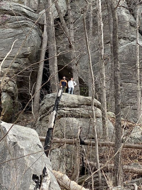 Two people stand on a large rock near a cave entrance, surrounded by bare trees and rocky terrain.