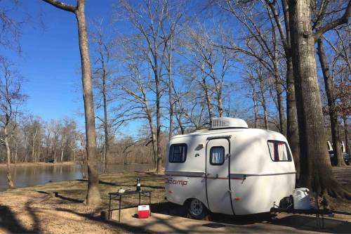 A small white camper parked by a serene pond, surrounded by bare trees under a clear blue sky.