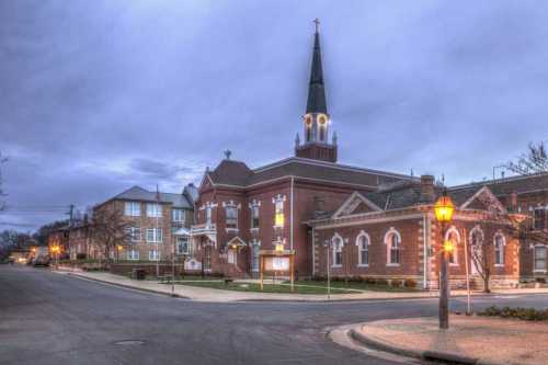 A historic church with a tall steeple, surrounded by buildings, under a cloudy evening sky. Street lamps illuminate the scene.