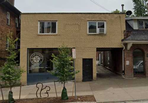 A two-story brick building with a storefront and an alleyway, surrounded by trees and parked cars.