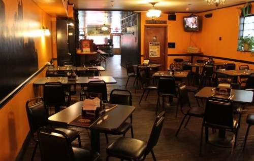 A cozy restaurant interior with empty tables and chairs, warm orange walls, and a TV in the background.