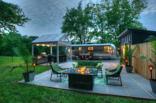 A cozy outdoor space featuring an Airstream trailer, patio furniture, and a grill surrounded by greenery.