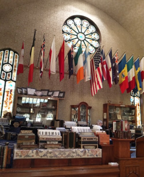 Interior of a room with a large stained glass window, featuring various flags hanging above a collection of accordions.