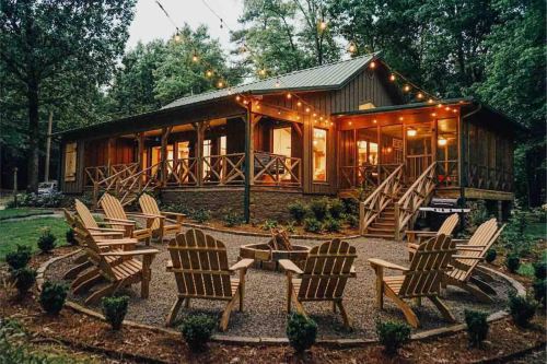 A cozy cabin surrounded by trees, featuring a fire pit and Adirondack chairs, illuminated by string lights at dusk.