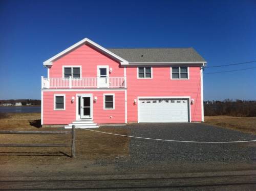 A bright pink two-story house with a gray roof, garage, and balcony, set against a clear blue sky.