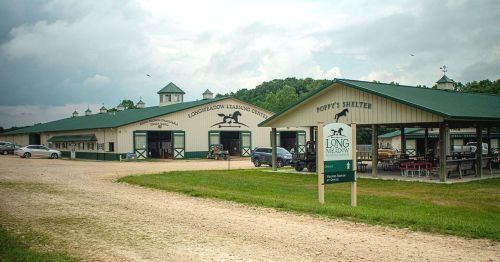 A horse shelter with multiple buildings and a sign, set in a grassy area under a cloudy sky.