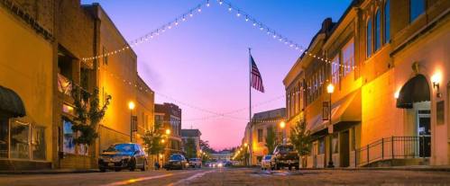 A charming street at dusk, lined with lights and trees, featuring an American flag and parked cars.