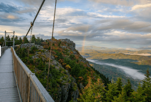 A scenic view from a suspension bridge overlooking mountains and valleys, with clouds and a rainbow in the sky.