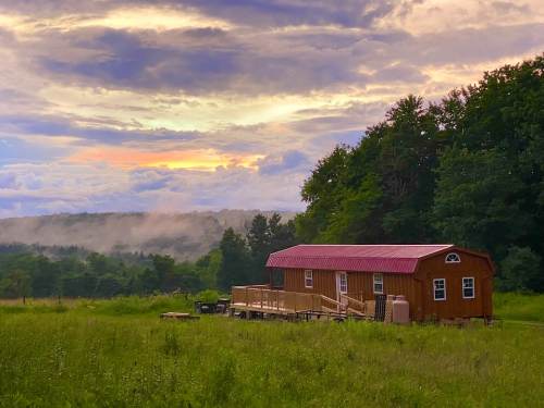 A rustic cabin with a red roof sits in a grassy field, surrounded by trees and a colorful sunset sky.