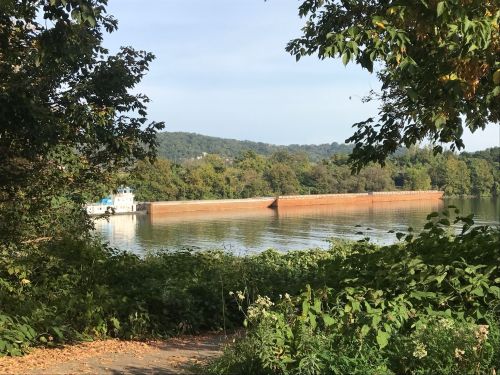 A serene river scene with a barge near a brick wall, surrounded by lush greenery and distant hills under a clear sky.