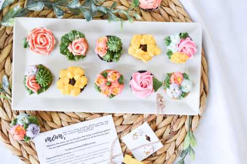 A platter of beautifully decorated cupcakes with floral designs, surrounded by greenery and a card.