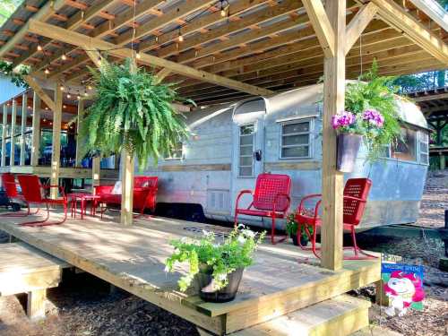 A vintage trailer under a wooden deck with hanging plants and red chairs, surrounded by greenery.