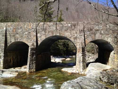 A stone bridge with three arches spans a clear stream, surrounded by trees and rocky terrain under a blue sky.