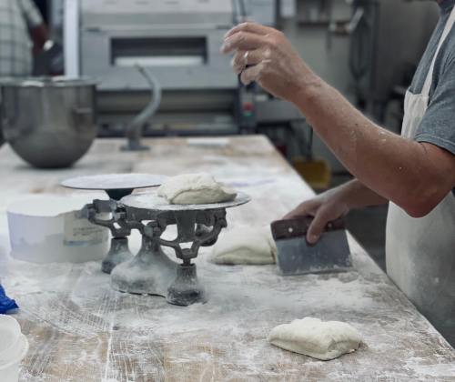 A baker weighs dough on a scale in a flour-dusted kitchen, with tools and ingredients visible in the background.