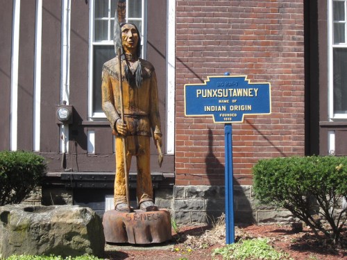 A wooden statue of a Native American figure stands next to a sign marking Punxsutawney's Indian origin.