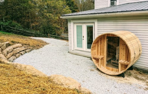 A wooden barrel sauna sits beside a gravel path leading to a house, surrounded by greenery and a stone wall.