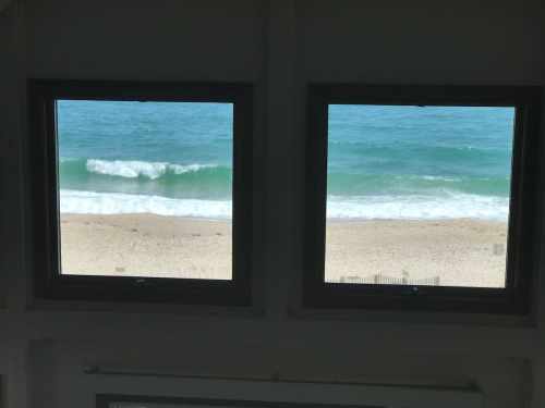Two windows framing a view of waves crashing on a sandy beach under a clear blue sky.
