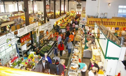 A bustling indoor market filled with shoppers, vendors, and colorful displays of food and flowers.