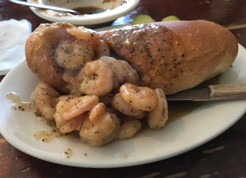 A shrimp po' boy sandwich with shrimp and sauce spilling out, served on a plate with a knife beside it.