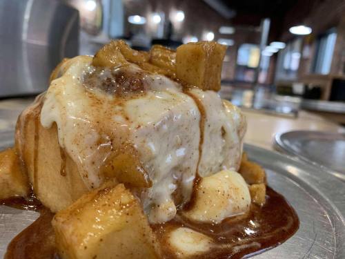 A close-up of a cinnamon roll topped with creamy icing and chunks of caramelized apples on a silver plate.
