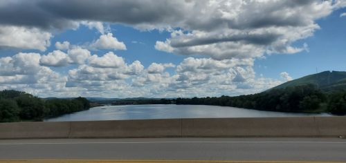 A scenic view of a river surrounded by lush greenery and hills, under a sky filled with fluffy clouds.