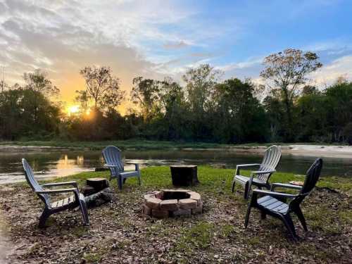 A serene lakeside scene with four chairs around a stone fire pit, framed by trees and a sunset sky.