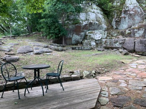 A small outdoor seating area with a table and two chairs, surrounded by rocks and greenery.