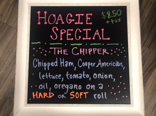 Chalkboard sign advertising a hoagie special: Chipped ham, cheese, veggies on a hard or soft roll for $8.50 + tax.
