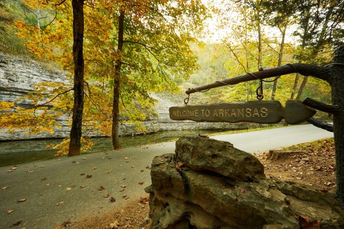 A wooden sign reading "Welcome to Arkansas" near a scenic road surrounded by autumn trees and rocky cliffs.