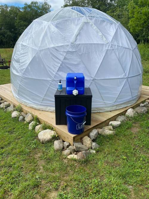 A geodesic dome on a wooden platform, with a blue water container and a black storage box in front, surrounded by rocks.