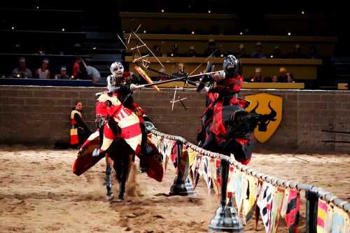 Two knights in armor charge at each other on horseback, wielding lances in a jousting match in a medieval arena.