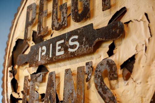 Close-up of a weathered sign reading "Peggy's Pies," featuring a rolling pin design and peeling paint.