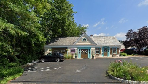 A small, charming building with a light-colored exterior, surrounded by greenery and a parking lot.