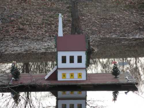 A small, whimsical house on a dock, surrounded by water and trees, reflecting in the calm surface.