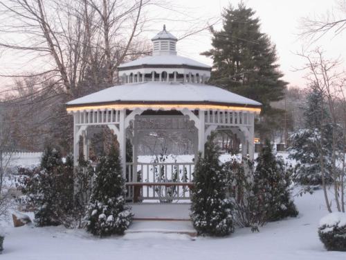 A snow-covered gazebo surrounded by evergreen shrubs, with a serene winter landscape in the background.