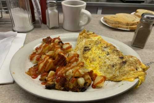 A plate of breakfast featuring an omelet, hash browns drizzled with ketchup, toast, and a coffee cup in the background.